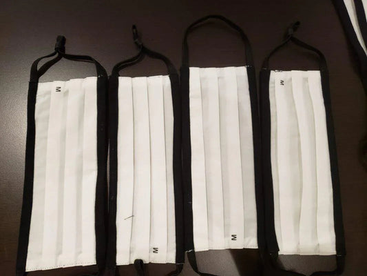 Four white pleated cotton face masks with black trim and ear loops on dark surface