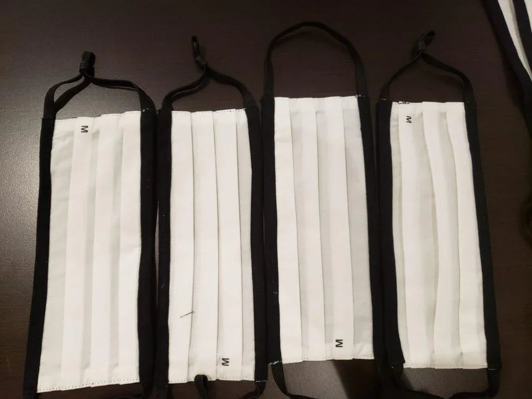 Four white pleated cotton face masks with black trim and ear loops on dark surface