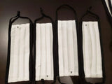 Four white pleated cotton face masks with black trim and ear loops on dark surface