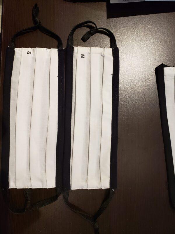 White pleated cotton face masks with black trim in small and medium sizes on dark surface