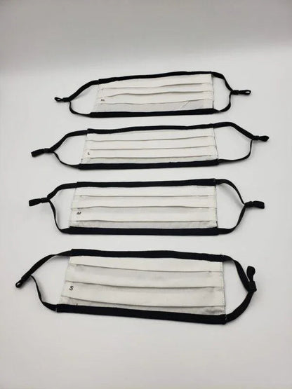 Four pleated white cotton face masks with black ear loops in various sizes on a white background.